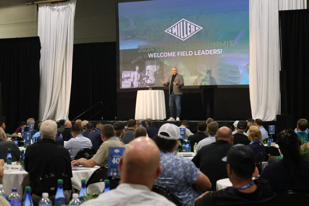 A speaker on a stage in front of people sitting at banquet tables.