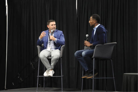 Two men sit on stools and talk with microphones